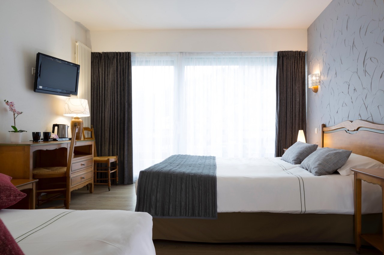 Chambre lumineuse avec lit double, bureau, télévision murale, rideaux sombres et grande fenêtre avec voilages.