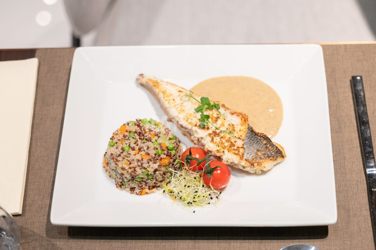 Filet de poisson grillé avec sauce, accompagné de quinoa, tomates cerises et pousses, servi sur une assiette blanche.