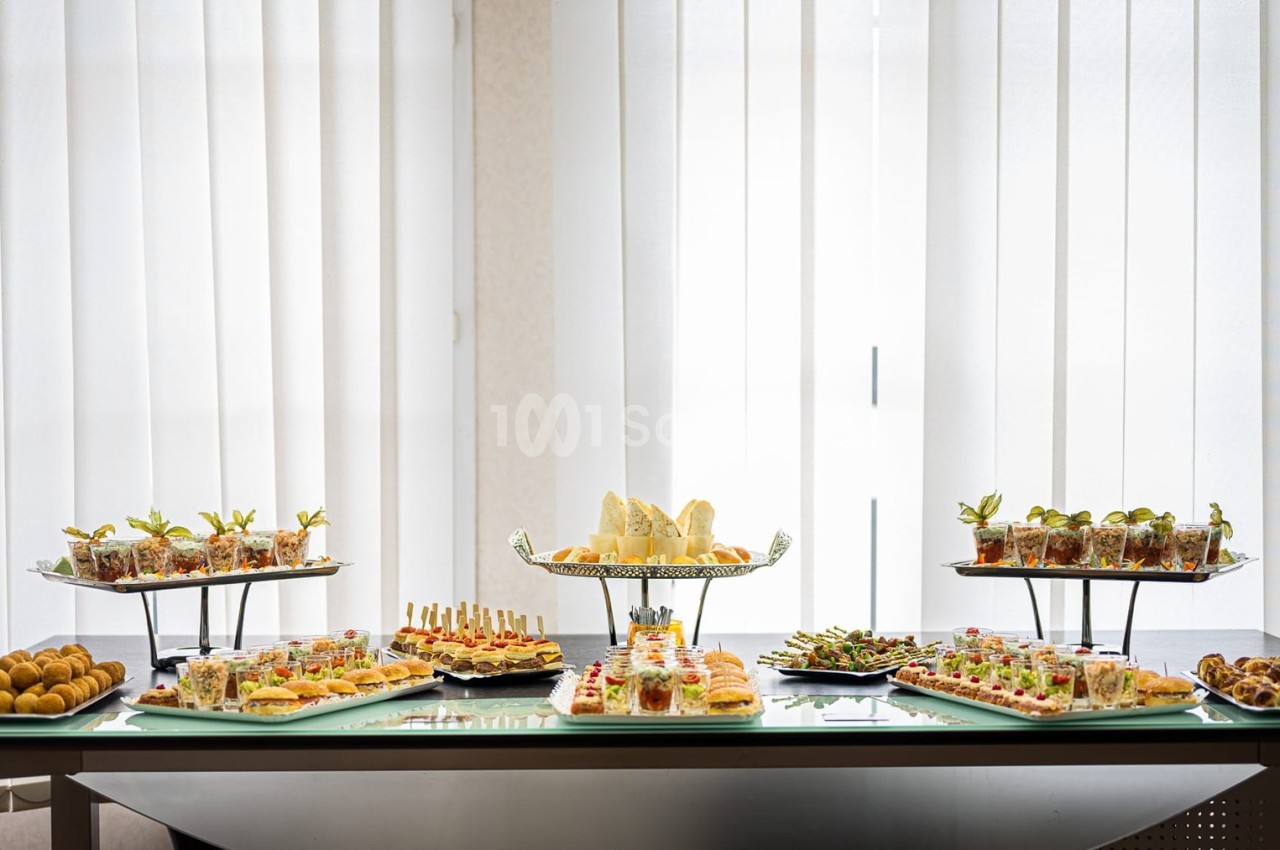 Buffet de table avec divers plats et amuse-bouches présentés sur des plateaux devant une grande fenêtre avec rideaux.