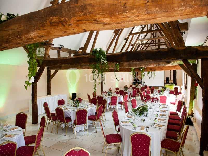 Salle de réception lumineuse avec poutres apparentes, tables rondes dressées et chaises rouges.