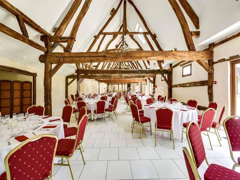 Salle de réception avec poutres apparentes, tables rondes dressées et chaises rouges disposées en rangées.