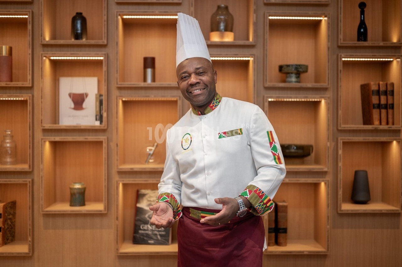 Un chef en tenue professionnelle blanche et rouge pose devant une étagère en bois décorée d'objets variés.