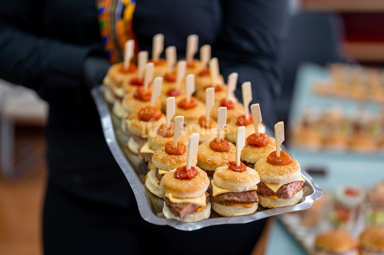 Plateau de mini-burgers garnis de sauce et maintenus par des piques en bois, tenu par une personne en tenue noire.