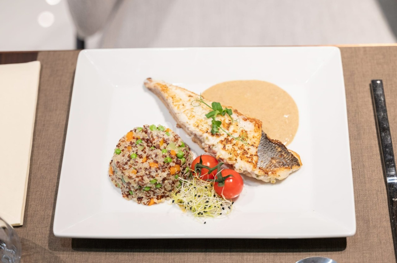 Filet de poisson grillé avec sauce, accompagné de quinoa, tomates cerises et pousses, servi sur une assiette blanche.