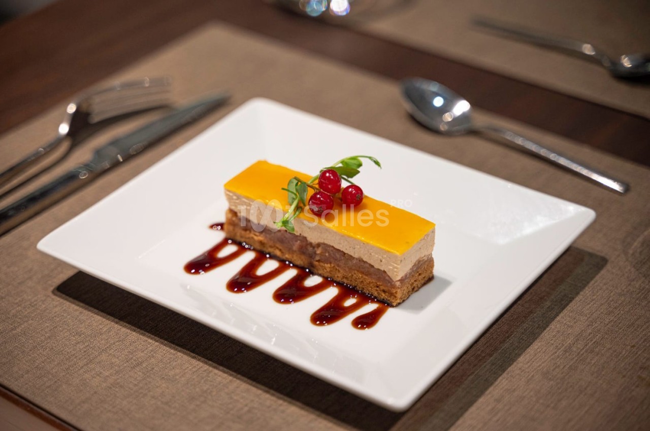 Dessert rectangulaire avec couches colorées, décoré de groseilles et servi sur un coulis rouge dans une assiette blanche.
