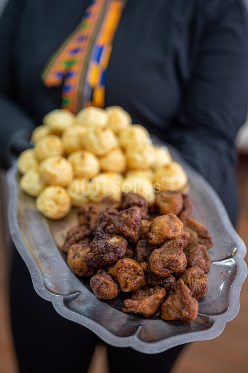 Plateau en métal contenant des beignets dorés et des boulettes frites, tenu par une personne en tenue sombre.