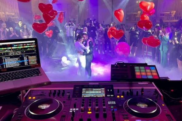 Vue depuis une table de mixage d'un DJ lors d'une fête avec un couple dansant entouré de ballons rouges en forme de cœur.