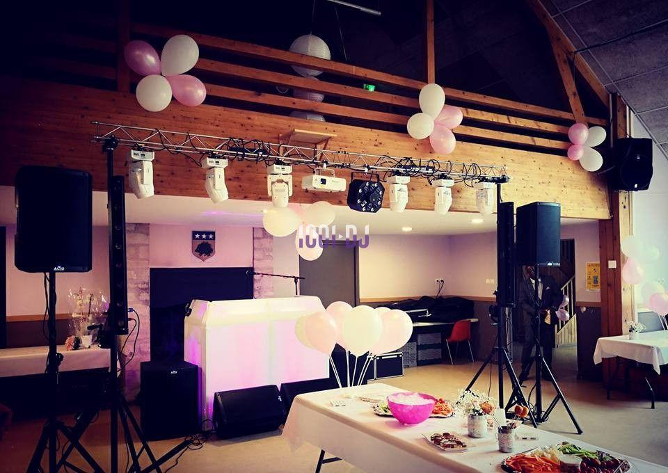 Salle décorée pour un événement avec ballons roses et blancs, tables dressées et matériel de sonorisation installé.