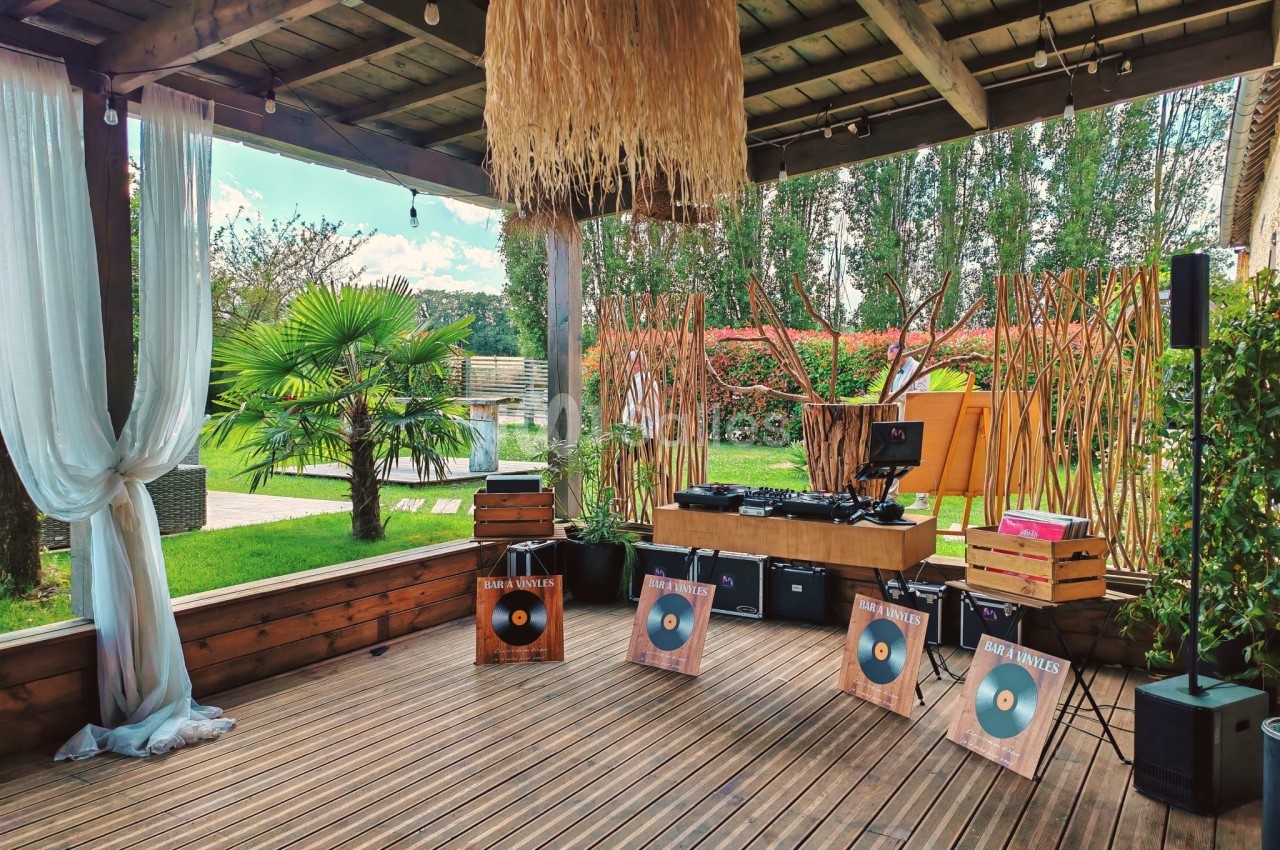 Espace extérieur couvert avec platines DJ, décor tropical, plantes, rideaux blancs et panneaux en bois.
