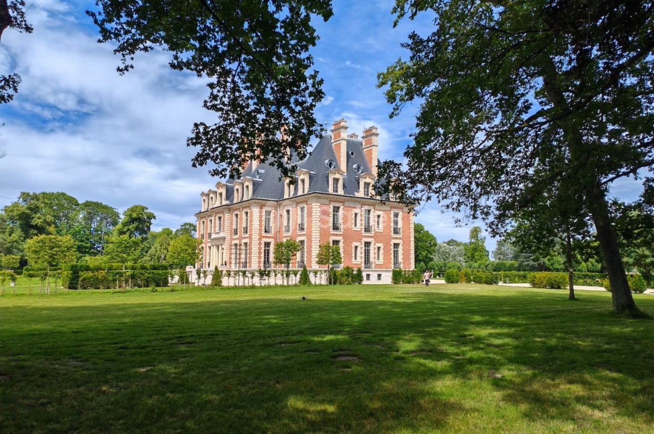 Manoir en briques rouges entouré d'un parc verdoyant, avec des arbres et un ciel partiellement nuageux.