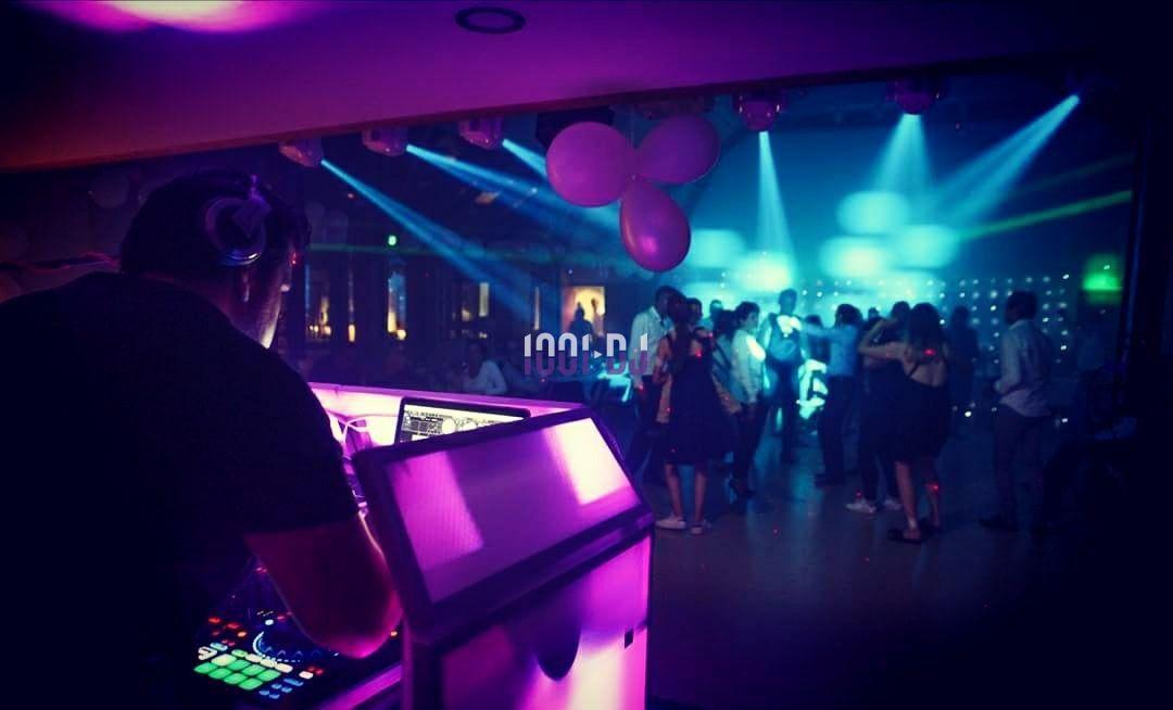 Un DJ mixe sur une console lumineuse dans une salle sombre avec des lumières colorées et des personnes dansant.