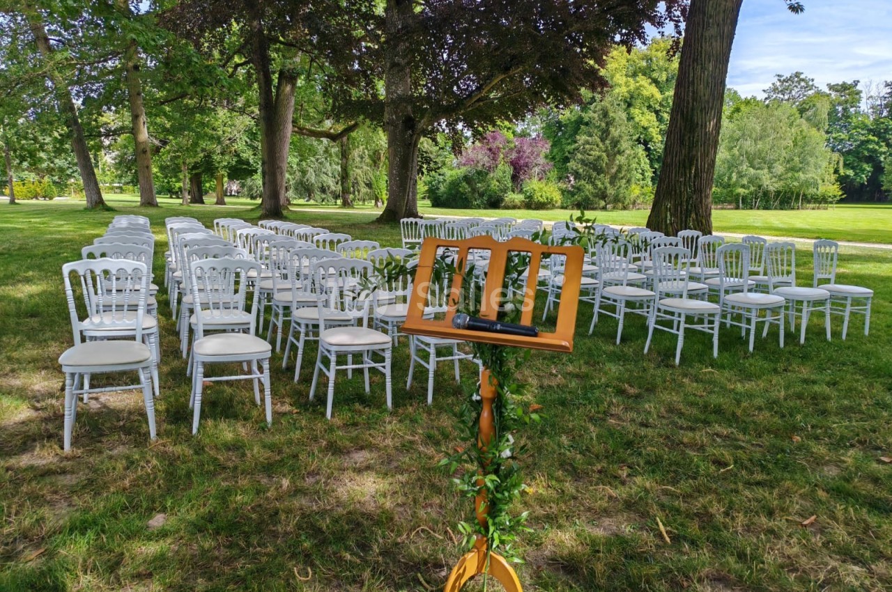 Chaises blanches alignées en extérieur sous des arbres, avec un pupitre en bois décoré de feuillage au premier plan.