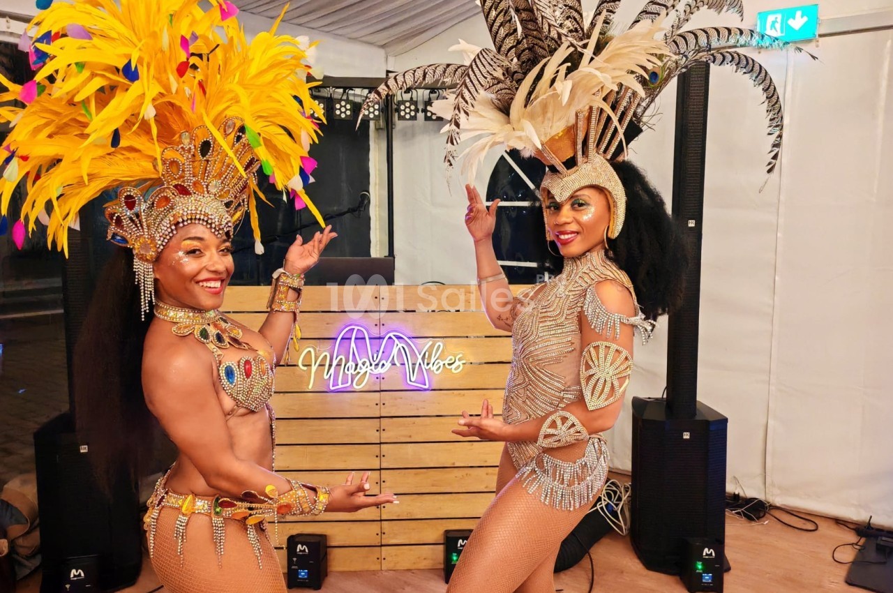 Deux danseuses en costumes traditionnels avec plumes colorées posent devant un panneau lumineux dans une tente.