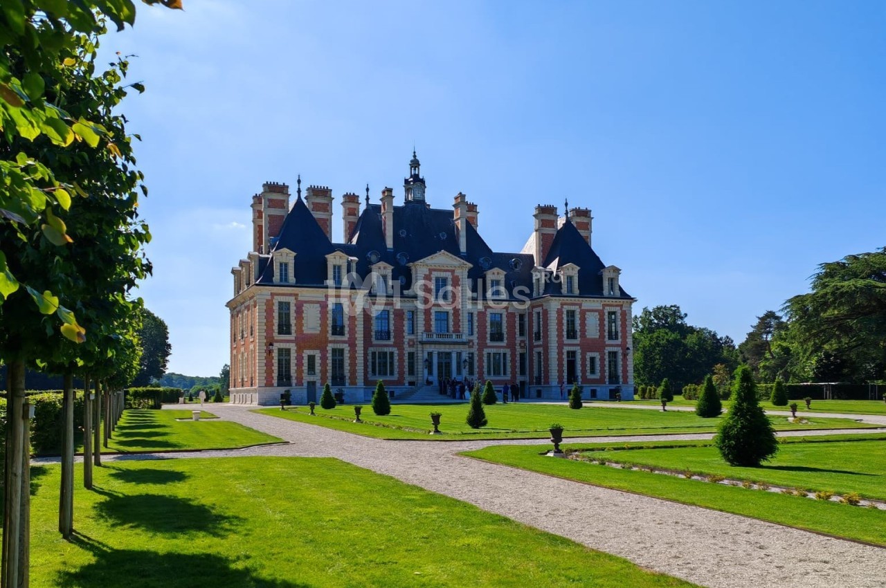 Château en briques et pierre entouré de jardins verdoyants sous un ciel bleu clair.