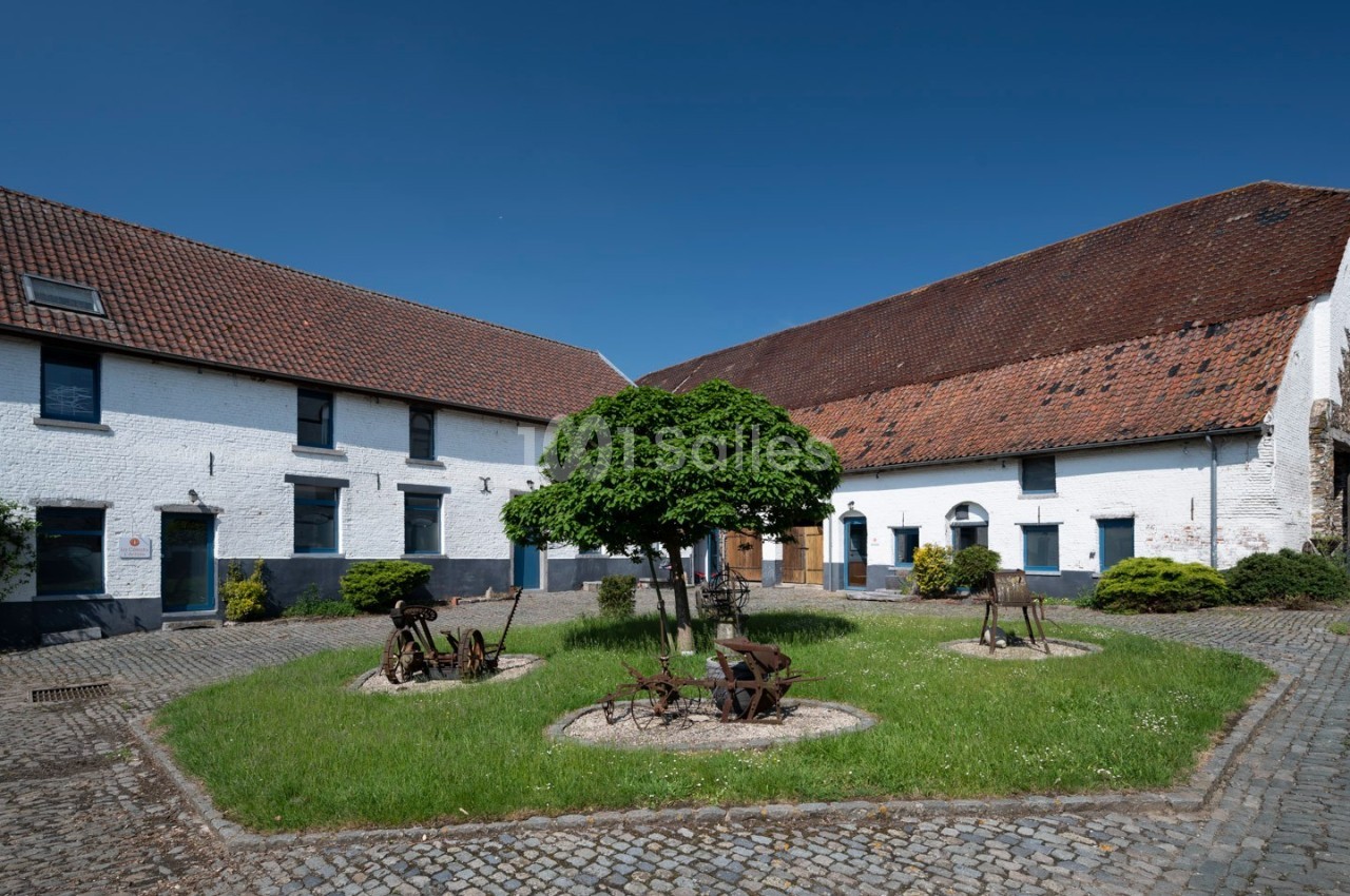 Cour intérieure pavée avec pelouse centrale, arbre, anciens outils agricoles et bâtiments en briques blanches.