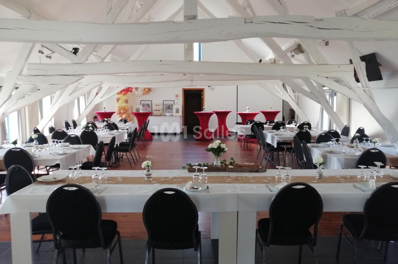 Salle lumineuse avec poutres apparentes, tables dressées pour un événement, chaises noires et décorations rouges.