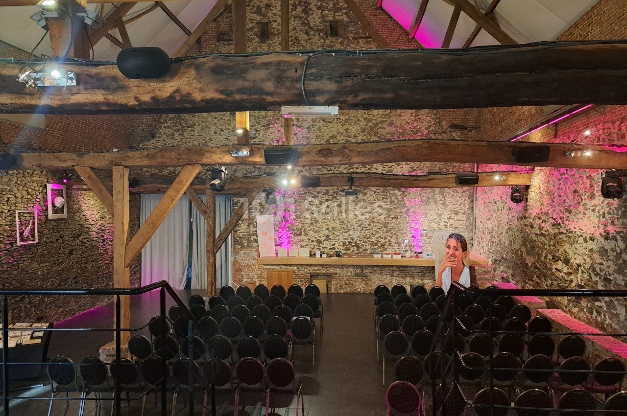 Salle en pierre avec poutres apparentes, rangées de chaises noires disposées face à une scène éclairée de lumières roses.