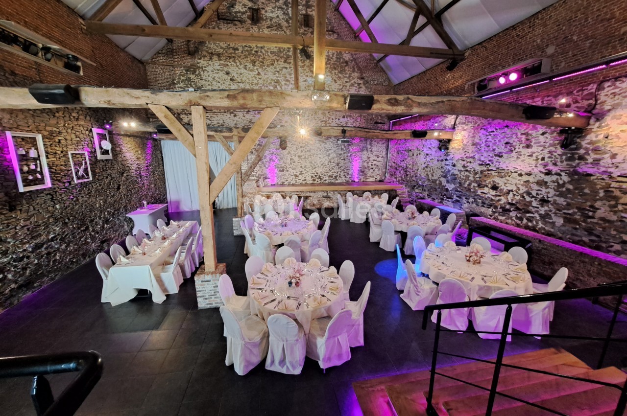 Salle de réception en pierre avec poutres apparentes, tables rondes dressées et éclairage violet.