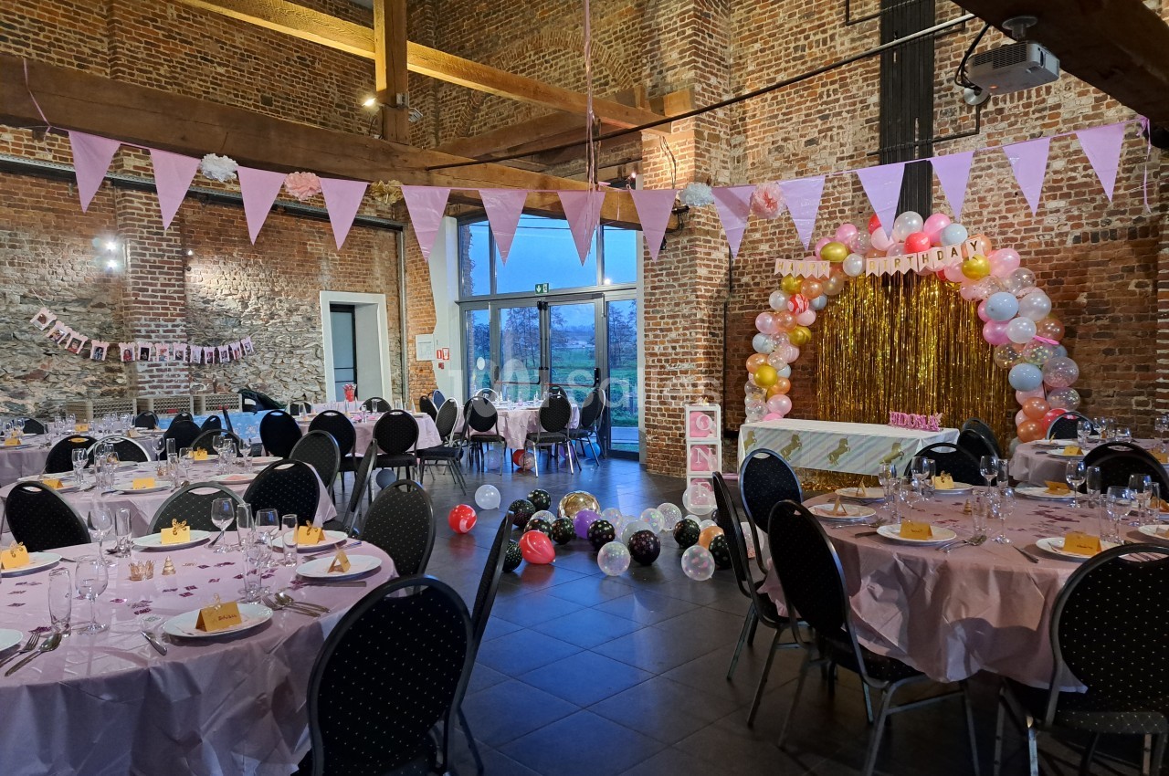 Salle décorée pour une fête avec tables dressées, guirlandes, ballons colorés et arche festive près d'un mur en briques.