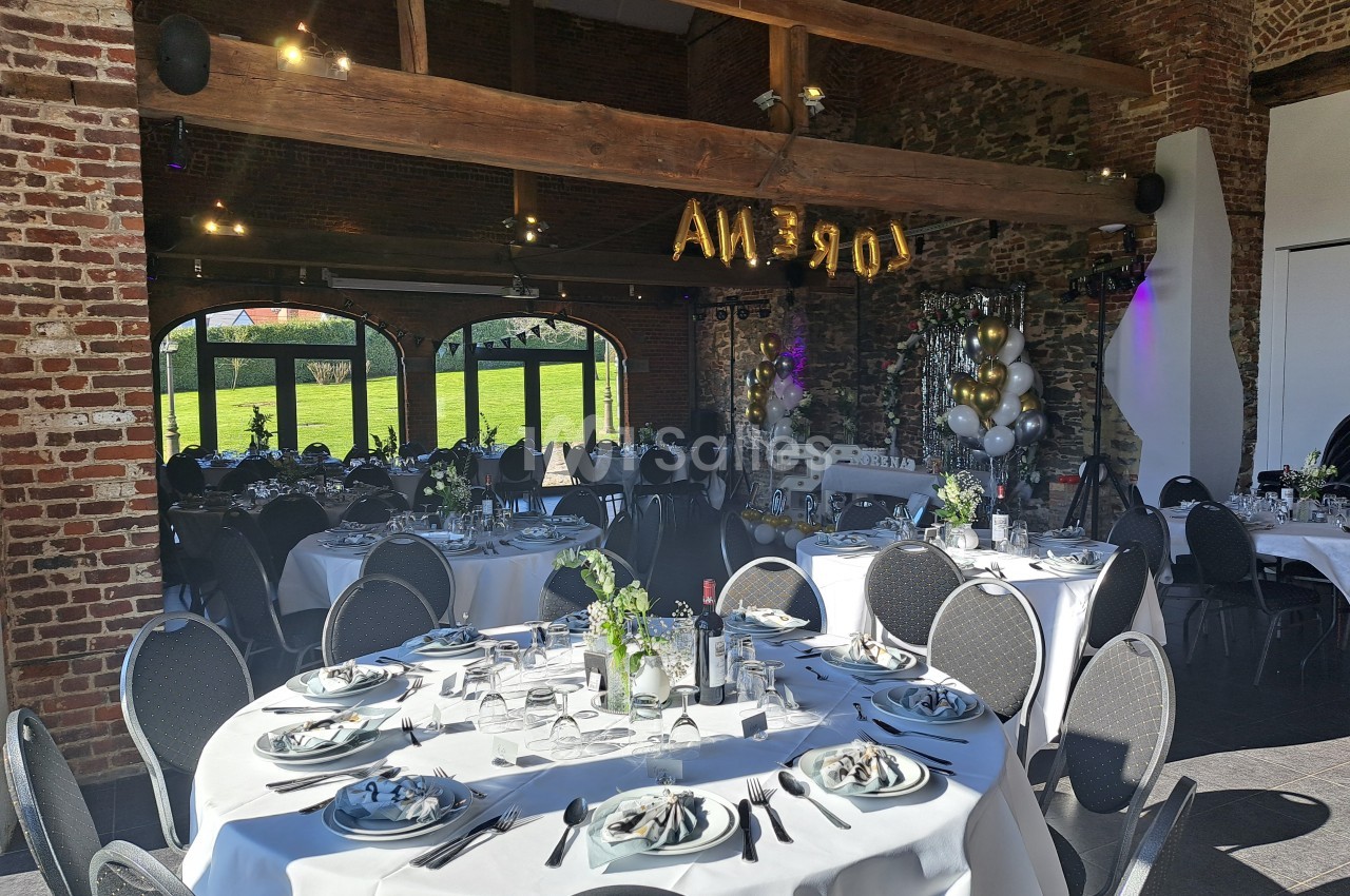 Salle de réception décorée avec des tables rondes dressées, des ballons et une vue sur un jardin à travers de grandes baies…