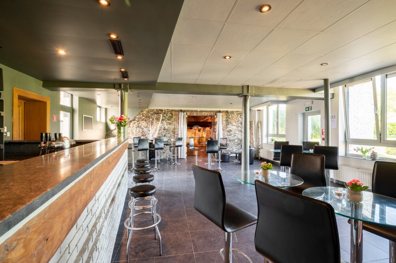 Salle lumineuse avec bar en bois, tables en verre, chaises noires et grandes fenêtres laissant entrer la lumière naturelle.