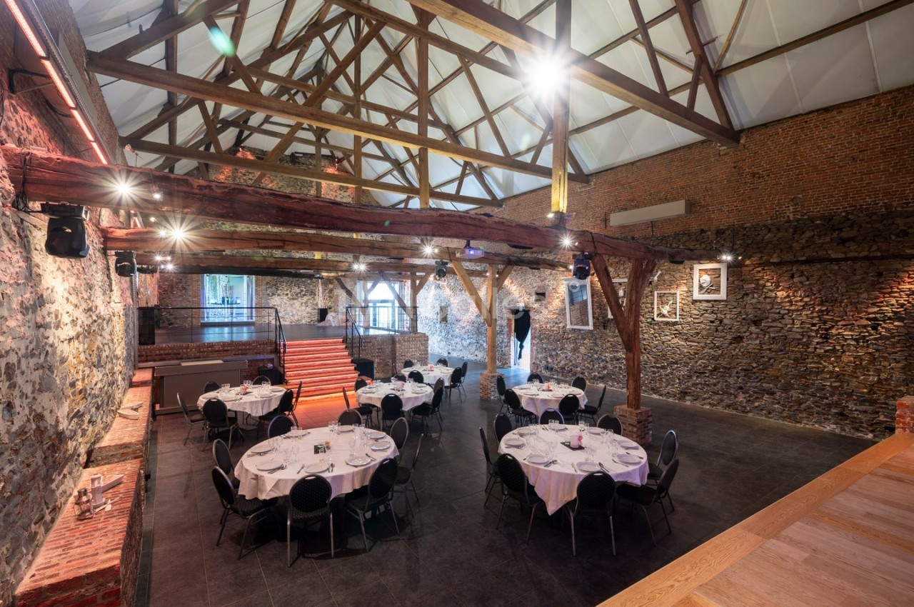 Salle de réception avec charpente en bois apparente, murs en pierre et tables rondes dressées pour un événement.