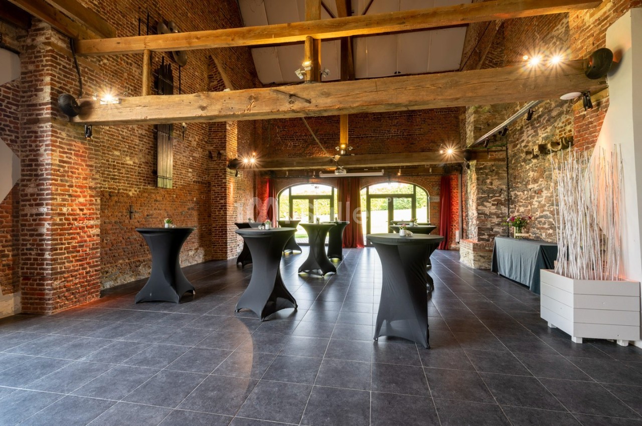 Salle en briques avec poutres apparentes, tables hautes noires et grandes ouvertures donnant sur un espace extérieur.