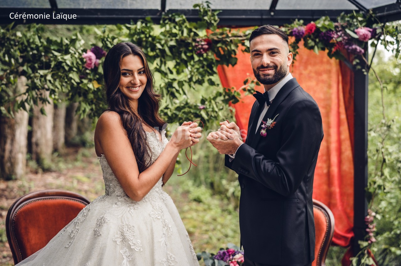 Un couple en tenue de mariage se tient les mains sous une arche décorée de feuillage et de fleurs.