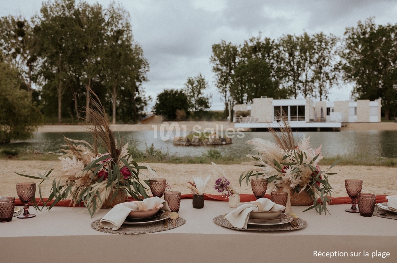 Table décorée avec des fleurs séchées et vaisselle élégante, installée en extérieur près d'un plan d'eau.