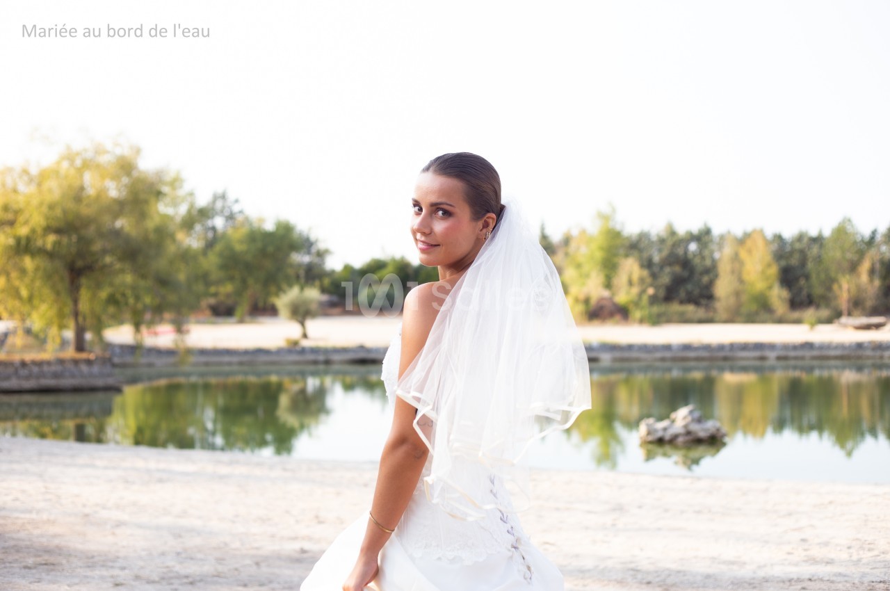 Femme en robe de mariée blanche avec un voile, posant près d'un plan d'eau entouré de végétation.