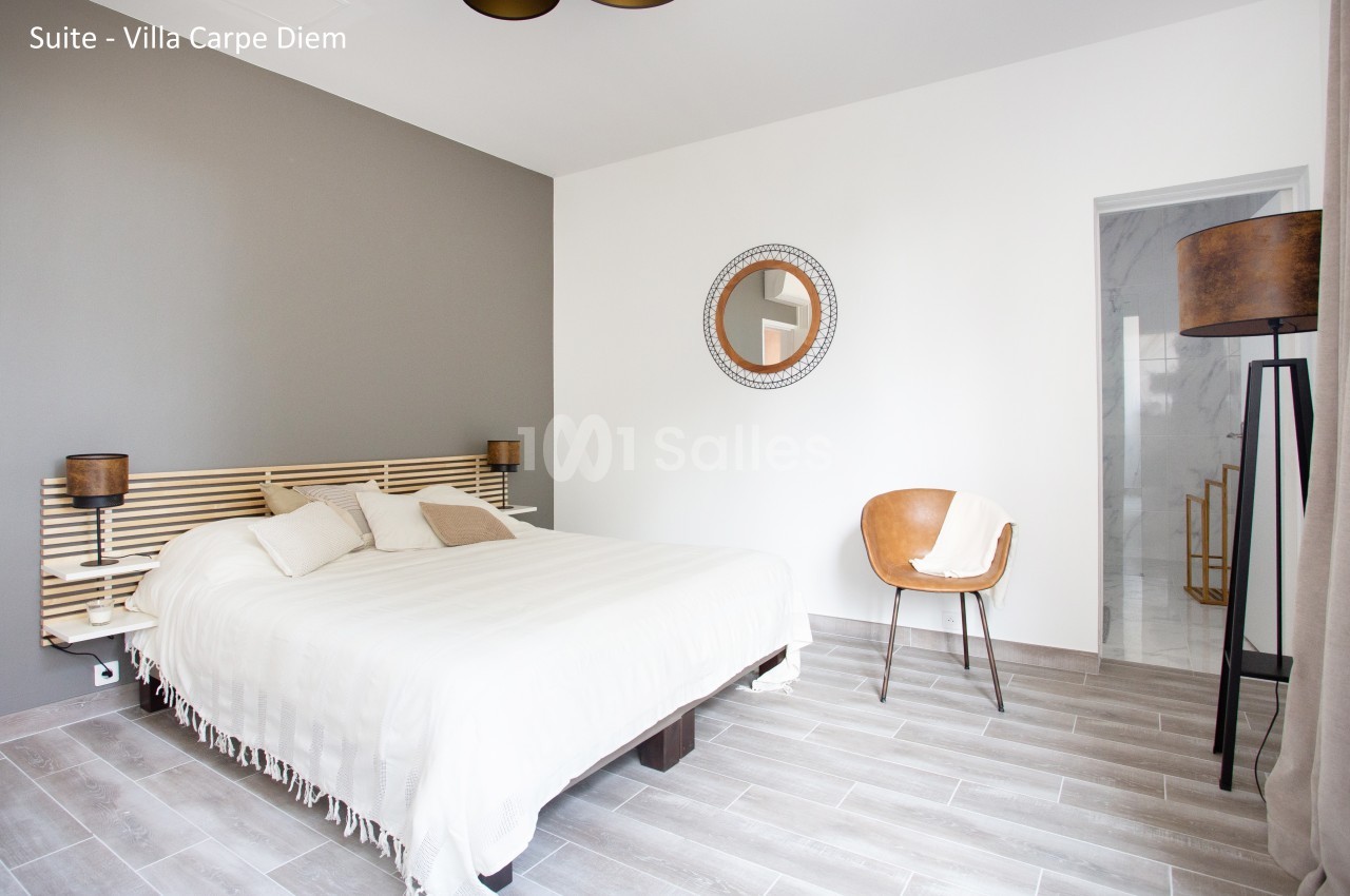 Chambre lumineuse avec lit double, mur gris, miroir rond, chaise en cuir et lampadaire sur sol en parquet clair.
