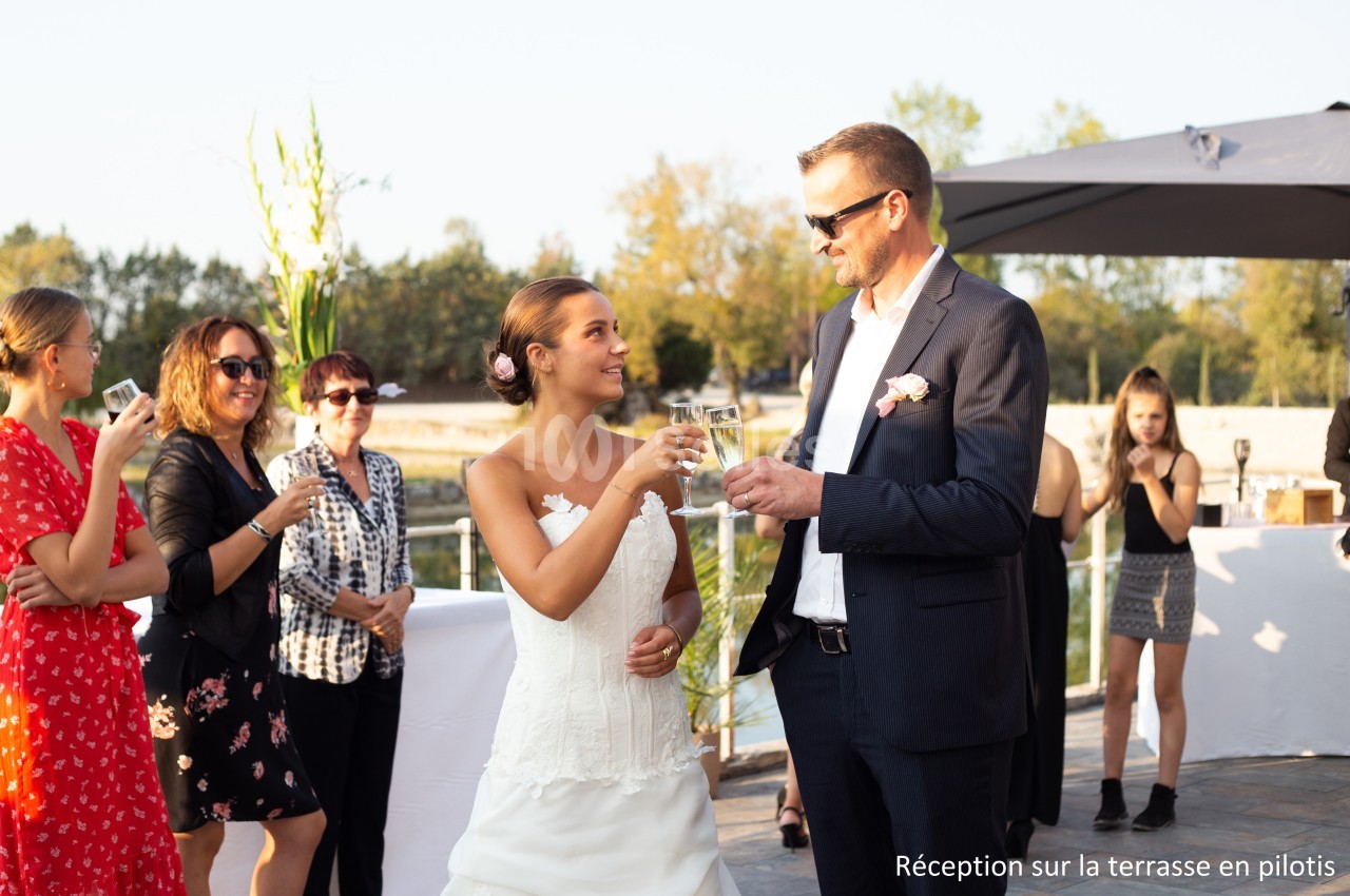 Une mariée et un homme en costume trinquent lors d'une réception en extérieur sur une terrasse ensoleillée.