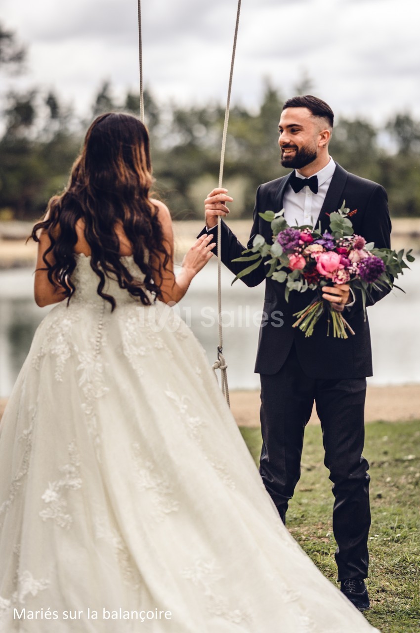 Un marié en costume tient un bouquet de fleurs face à une mariée en robe blanche près d'une balançoire en extérieur.
