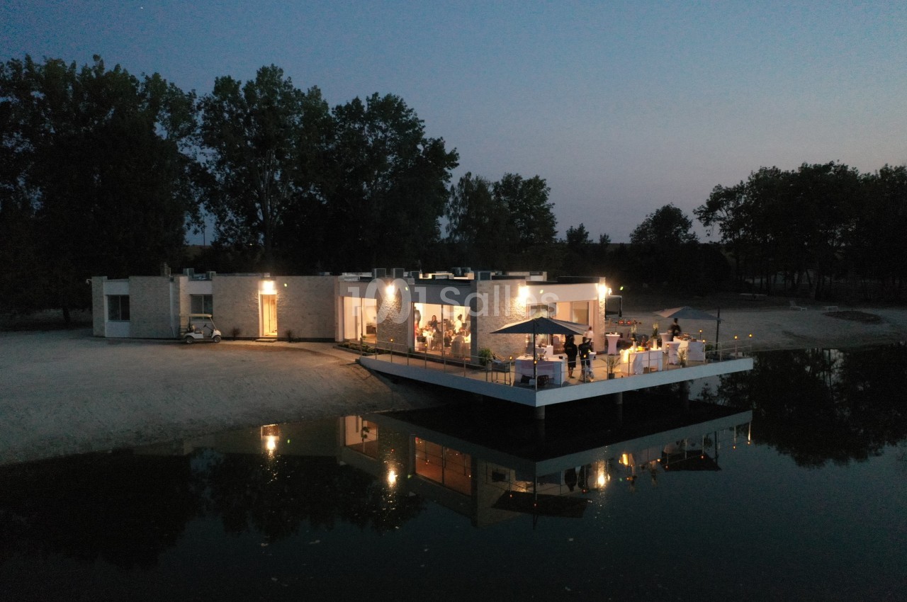 Vue aérienne d'une maison moderne au bord d'un plan d'eau, éclairée en soirée avec une terrasse animée.