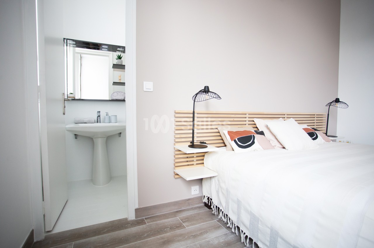 Chambre lumineuse avec lit double, tête de lit en bois clair, lampes noires et salle de bain attenante visible.