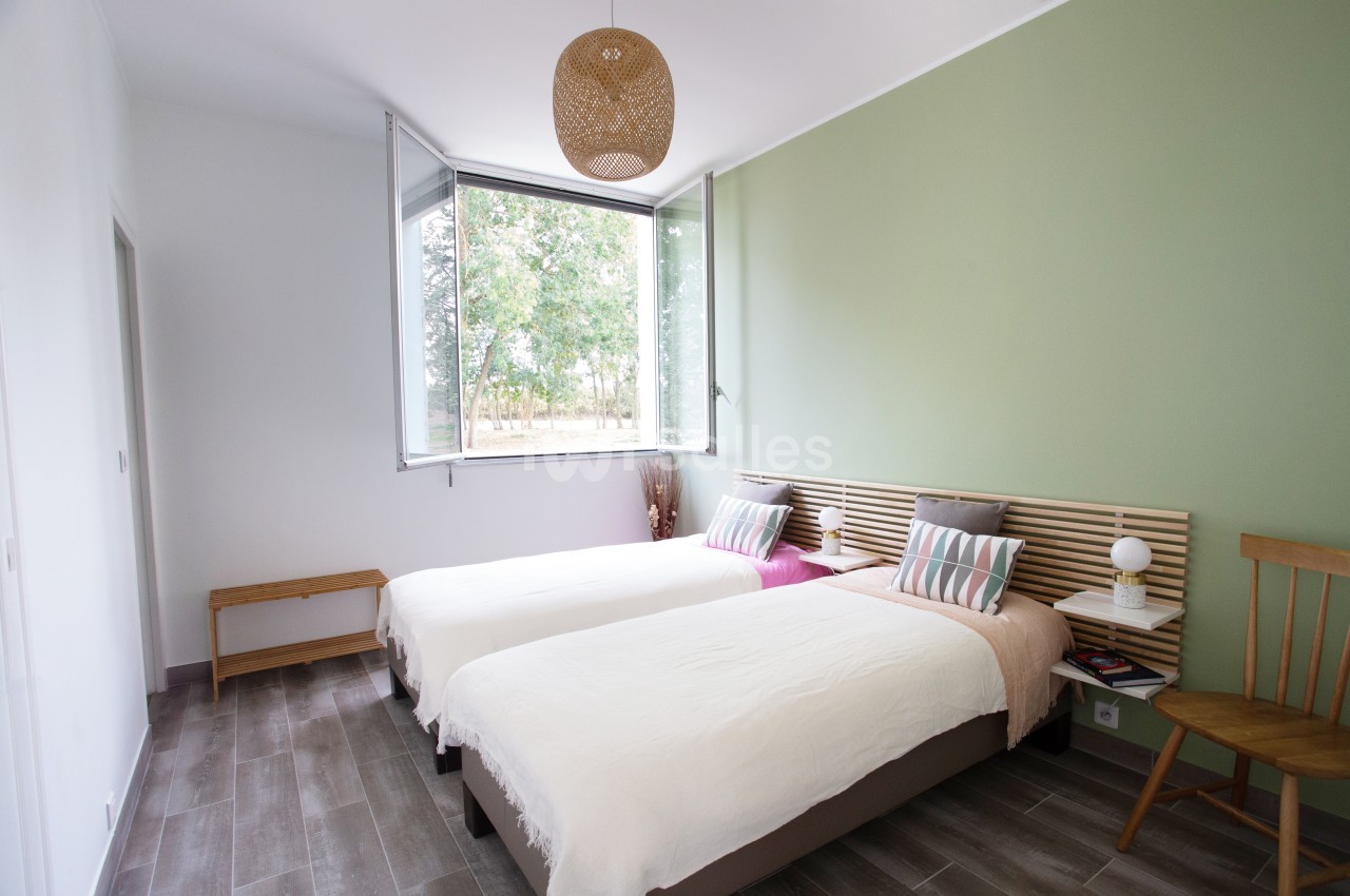 Chambre lumineuse avec deux lits simples, murs verts et blancs, parquet gris, fenêtre ouverte sur un jardin.