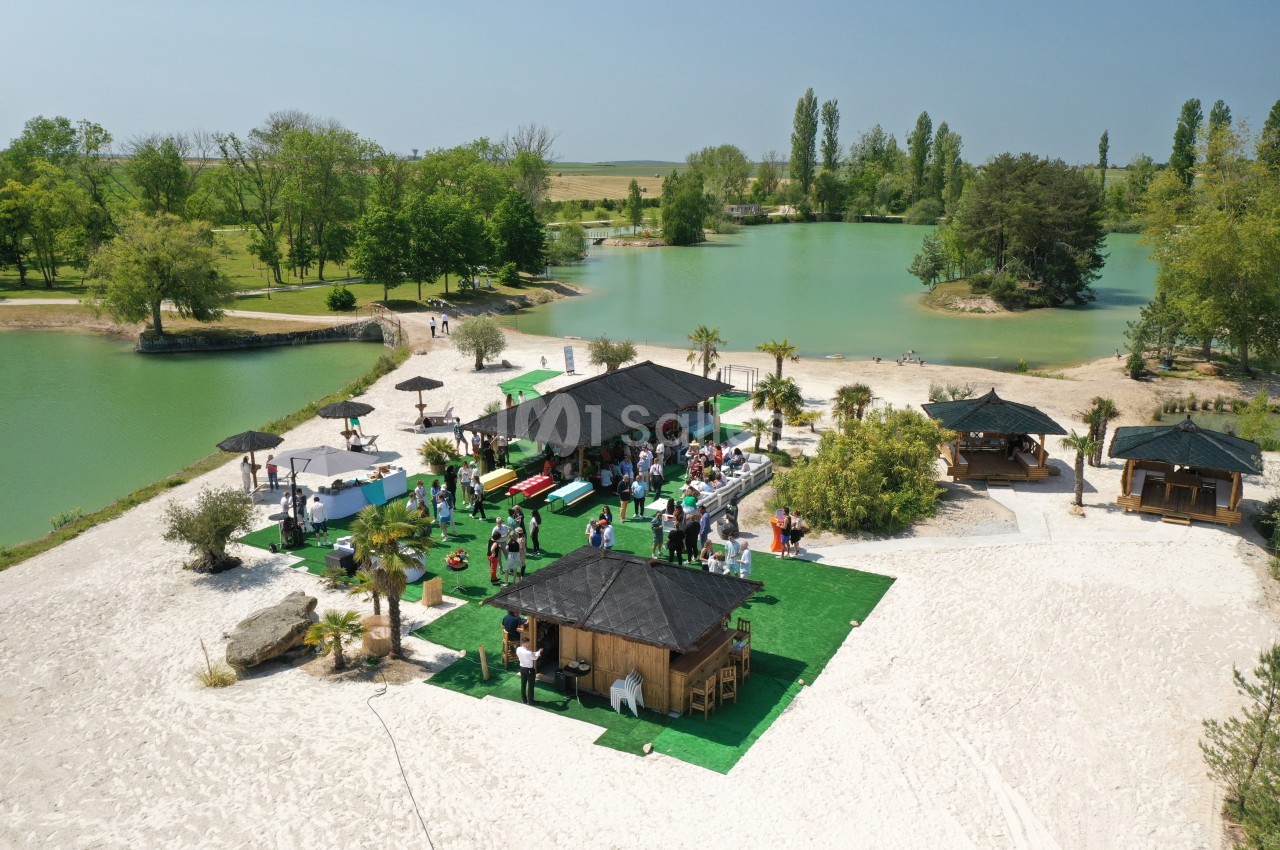 Vue aérienne d'une plage aménagée avec des kiosques, des parasols et des visiteurs près d'un lac entouré de verdure.