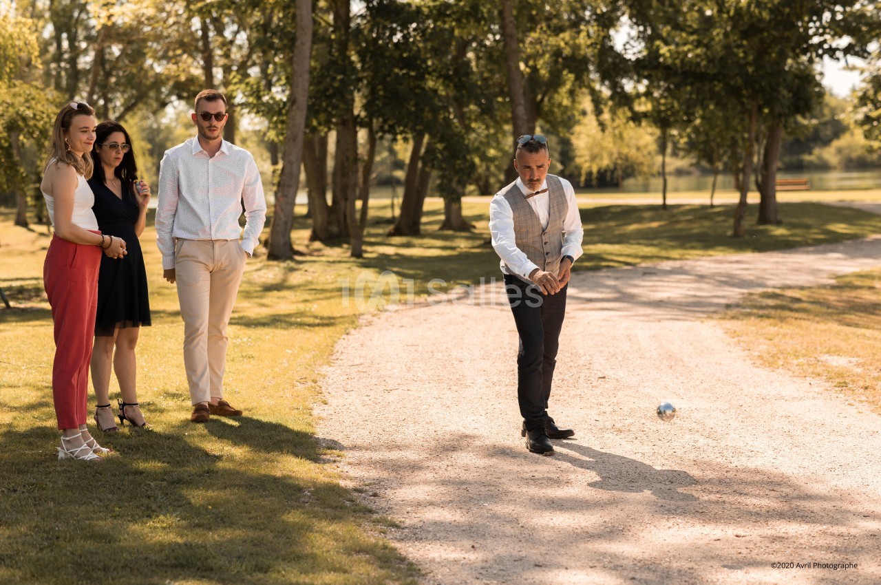 Un homme joue à la pétanque sur un chemin en gravier, observé par quatre personnes debout à l'ombre des arbres.