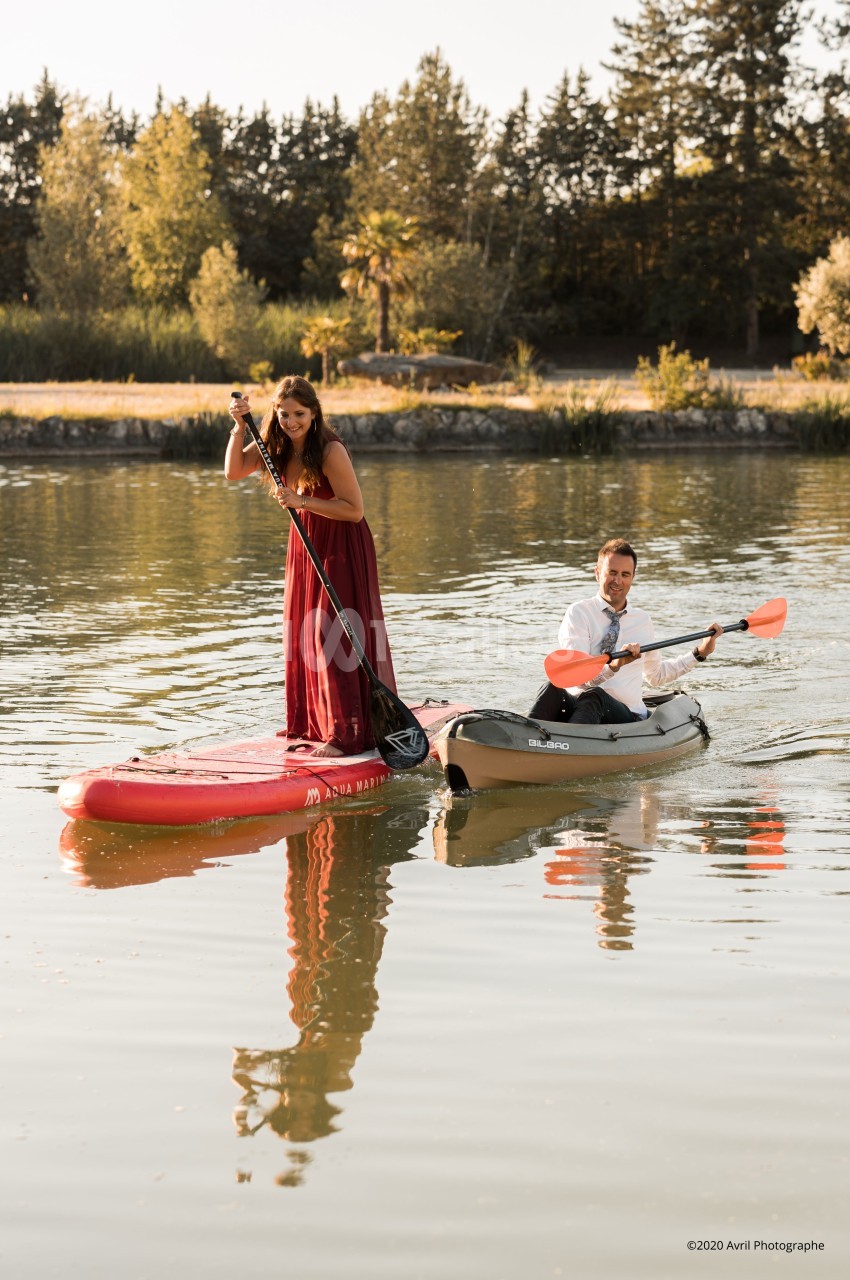 Une femme en robe rouge sur une planche de paddle et un homme en kayak pagaient sur un lac entouré d'arbres.