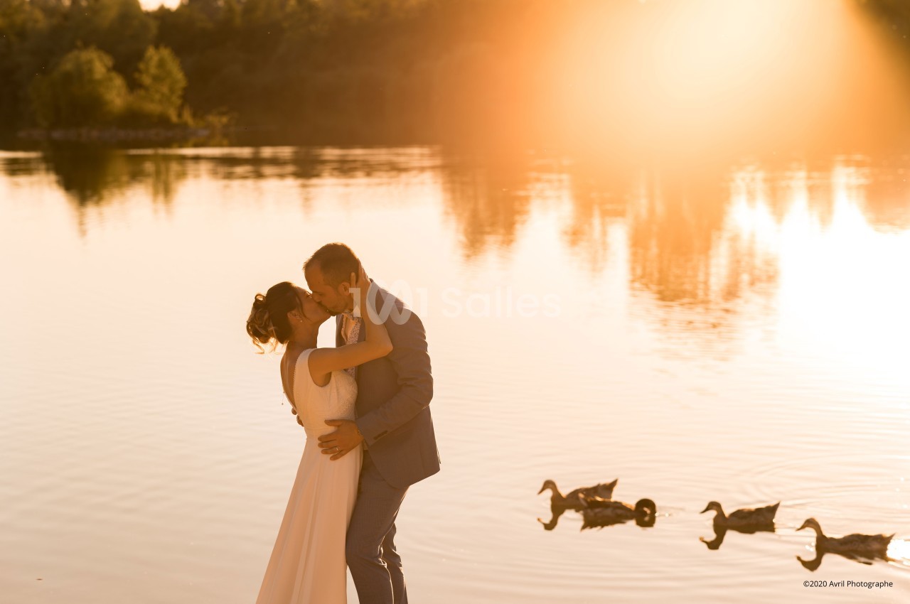 Un couple s'embrasse au bord d'un lac au coucher du soleil, avec des canards nageant sur l'eau calme.