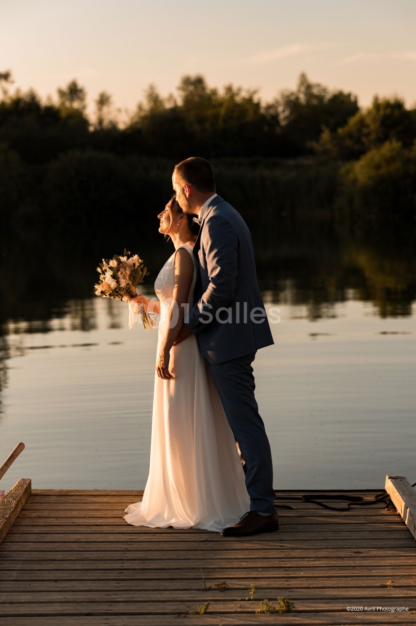 Un couple en tenue de mariage se tient sur un ponton en bois au bord d'un lac, éclairé par une lumière dorée.