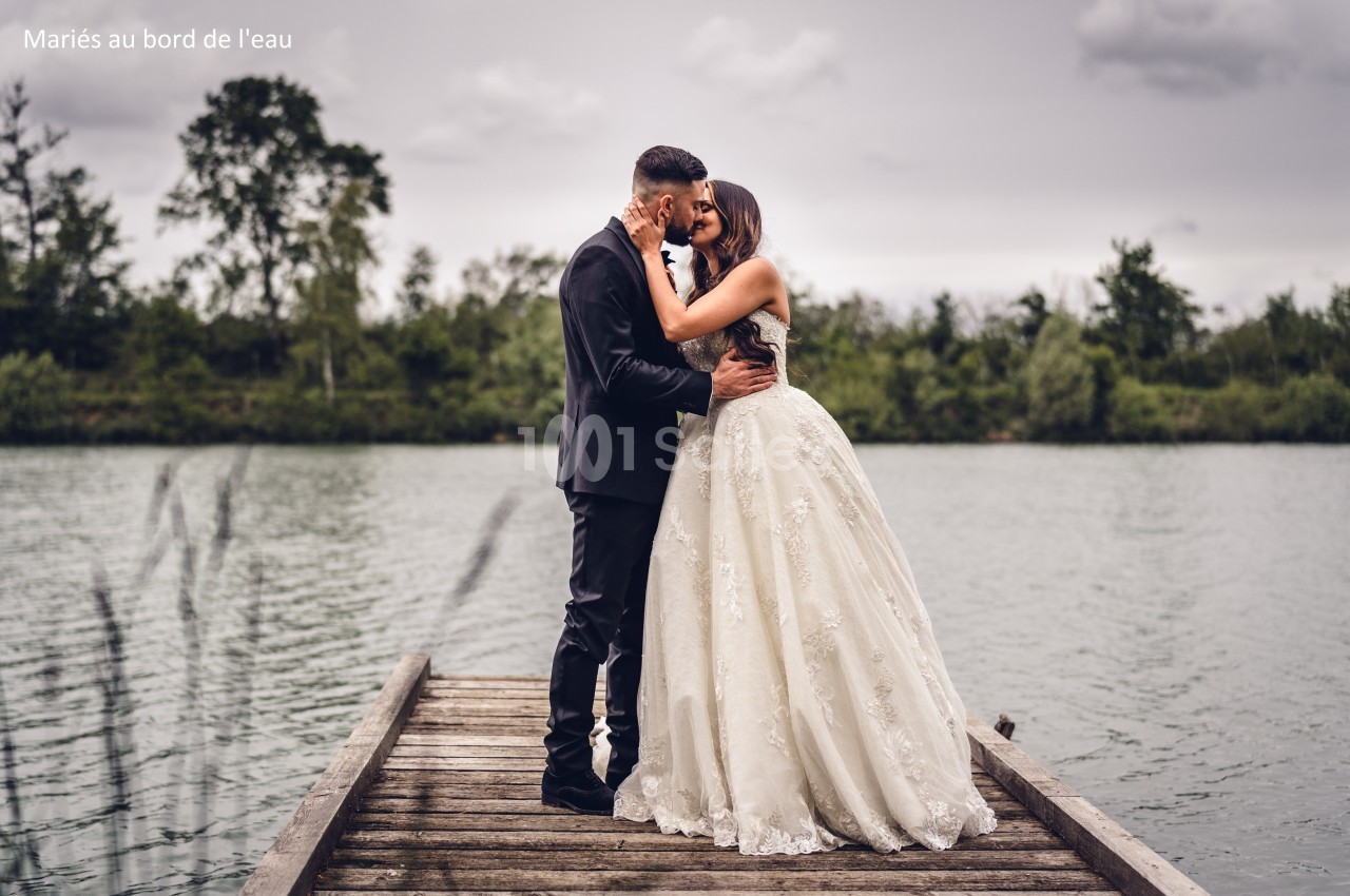 Un couple de mariés s'embrassant sur un ponton en bois au bord d'un lac, sous un ciel nuageux.
