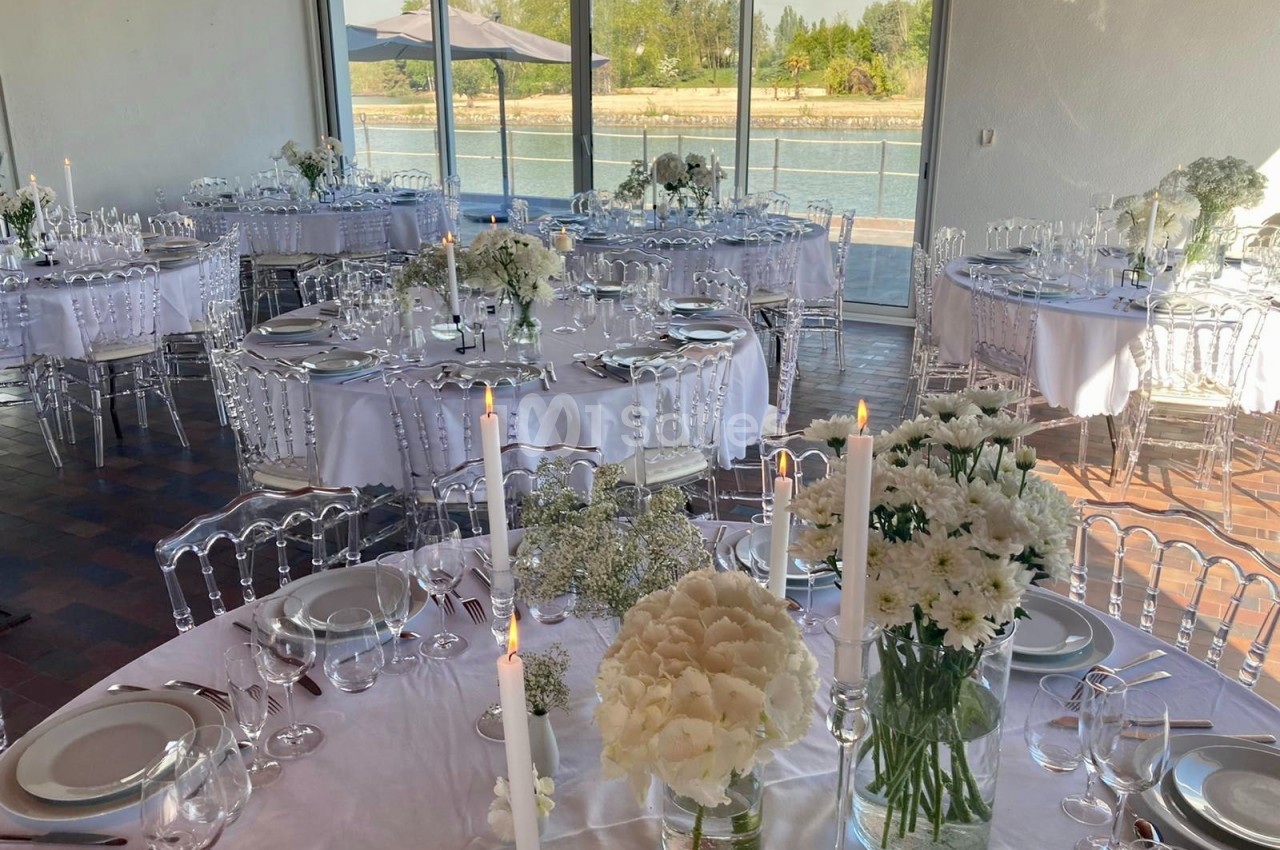 Salle de réception lumineuse avec tables rondes dressées, décorées de fleurs blanches et bougies, vue sur un plan d'eau.