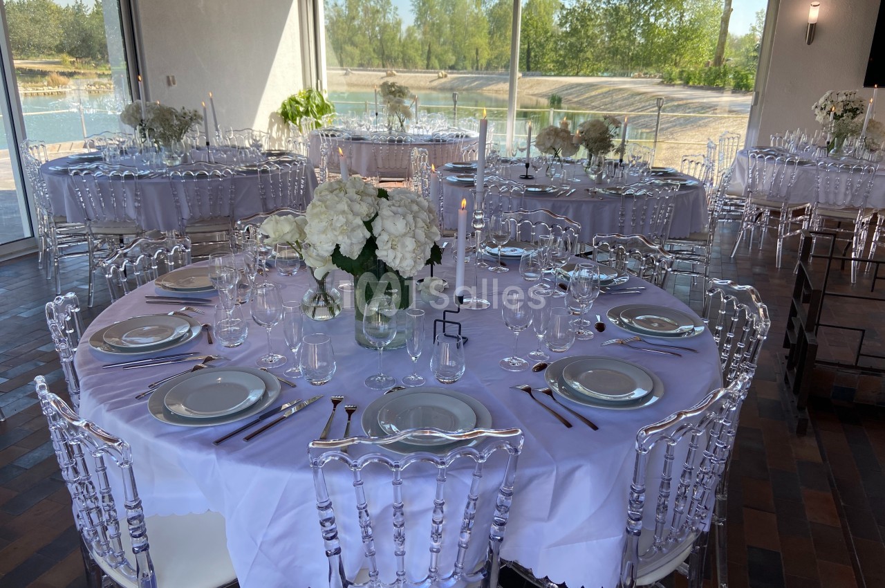 Tables rondes dressées avec nappes blanches, vaisselle élégante et bouquets de fleurs blanches dans une salle lumineuse.