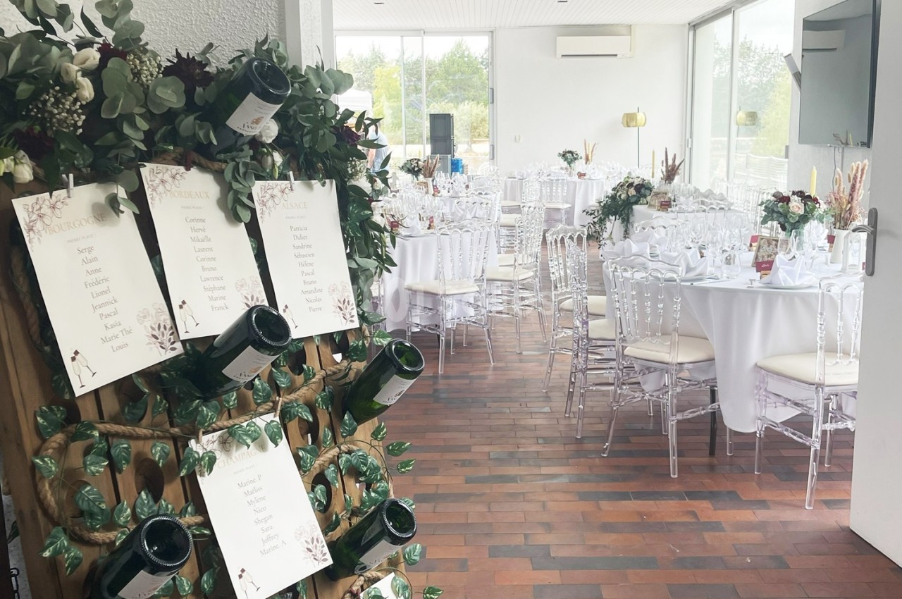 Salle de réception décorée pour un mariage avec tables dressées et plan de table affiché sur un support en bois.