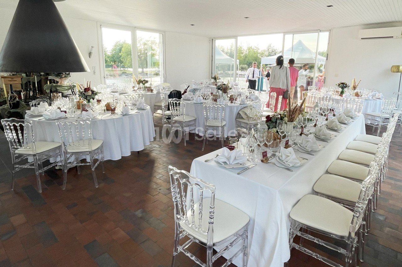 Salle de réception lumineuse avec tables dressées, chaises transparentes et vue sur l'extérieur.