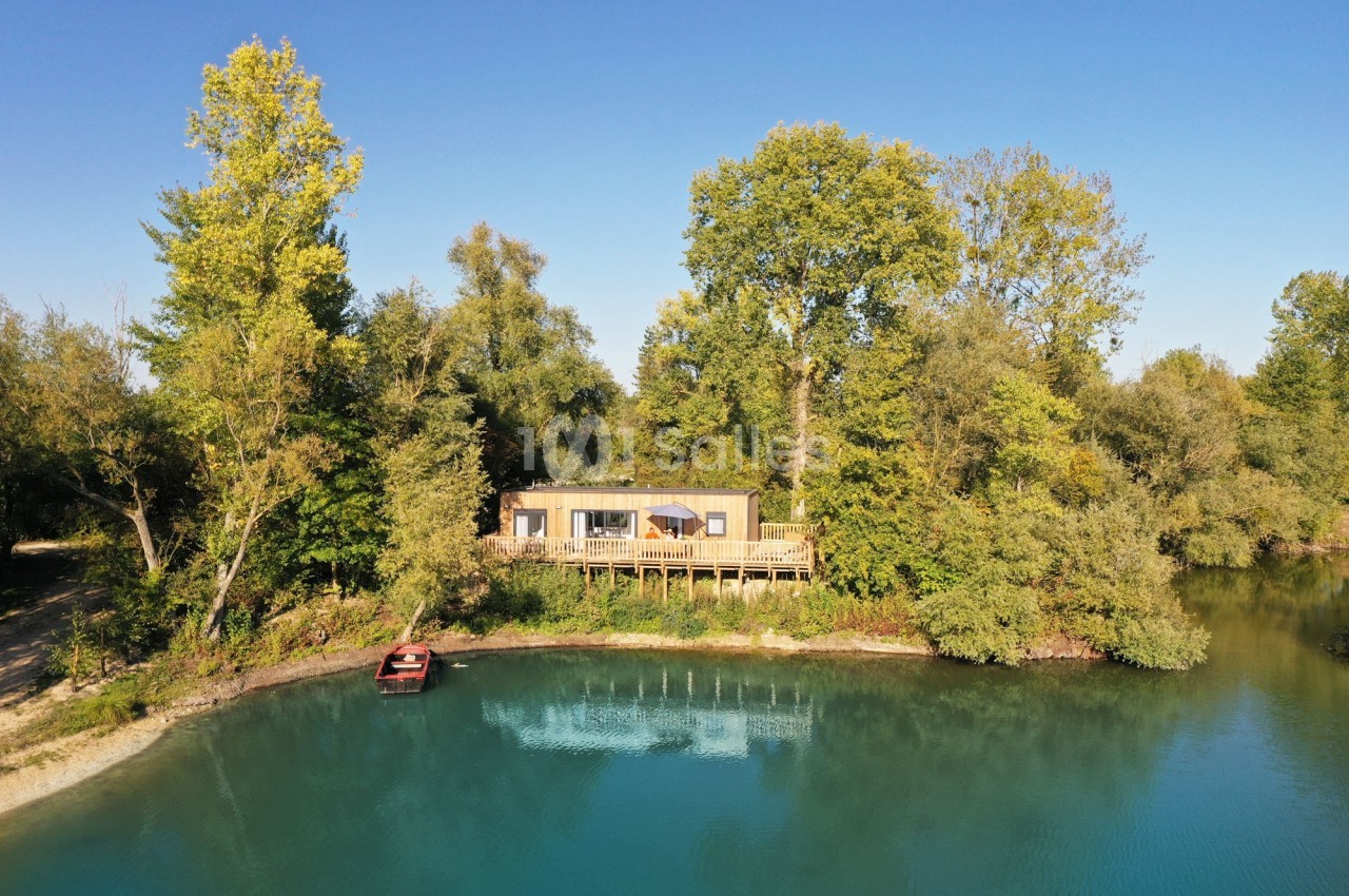 Maison en bois sur pilotis entourée d'arbres, située au bord d'un lac avec une eau turquoise et un petit bateau rouge.