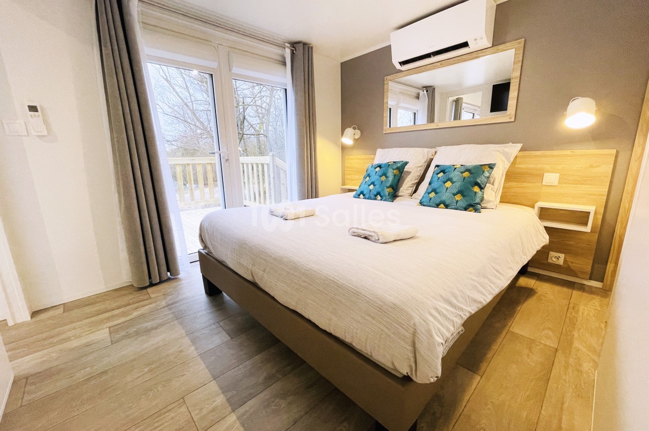 Chambre lumineuse avec lit double, coussins décoratifs, draps pliés, grande baie vitrée et sol en bois clair.