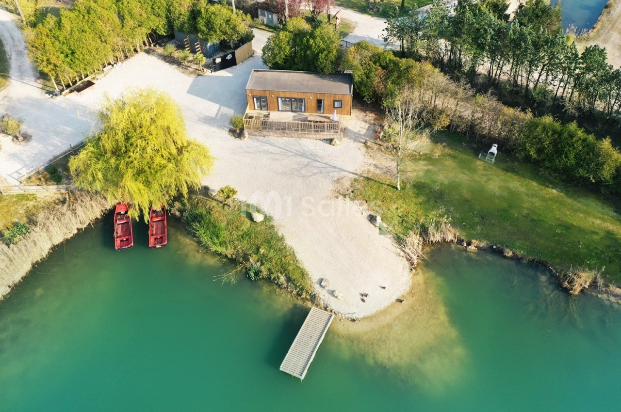 Vue aérienne d'une maison en bois près d'un lac avec un ponton, deux barques rouges et un espace arboré.