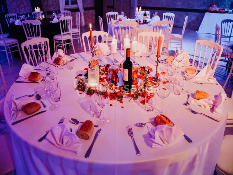 Table ronde élégamment dressée avec nappes blanches, bougies, pétales rouges, pain et bouteille de vin au centre.